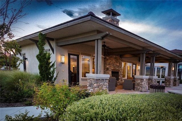 rear view of house with stucco siding, an outdoor stone fireplace, stone siding, a ceiling fan, and a patio area