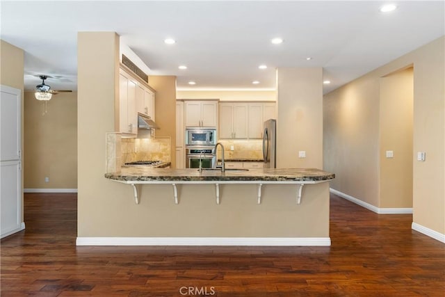 kitchen featuring a peninsula, dark stone counters, appliances with stainless steel finishes, under cabinet range hood, and a breakfast bar area