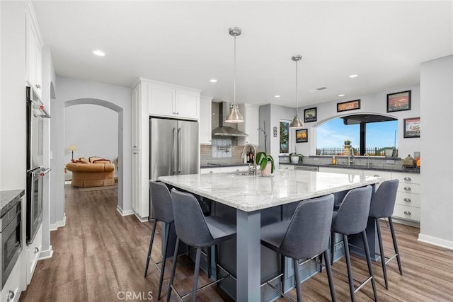 kitchen featuring arched walkways, wall chimney exhaust hood, dark wood finished floors, white cabinetry, and stainless steel appliances