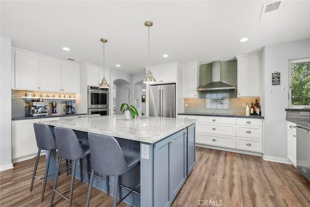 kitchen featuring white cabinets, arched walkways, wall chimney range hood, stainless steel appliances, and backsplash