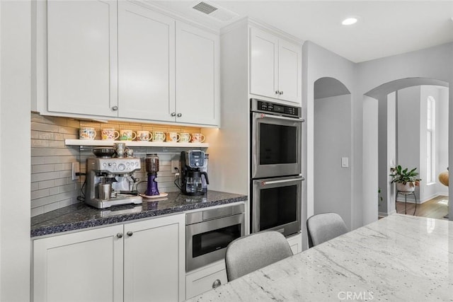 kitchen with white cabinetry, backsplash, dark stone counters, stainless steel appliances, and arched walkways