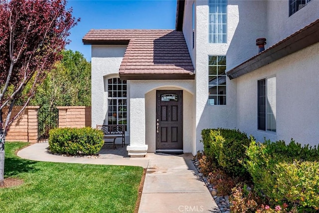 view of exterior entry with a yard, stucco siding, a tiled roof, and fence