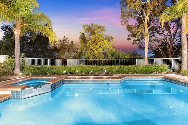 pool at dusk featuring a pool with connected hot tub and fence