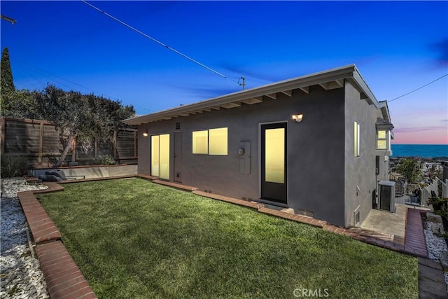back of house at dusk featuring central AC unit, stucco siding, fence, and a lawn