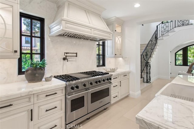kitchen featuring glass insert cabinets, light stone counters, decorative backsplash, custom range hood, and range with two ovens