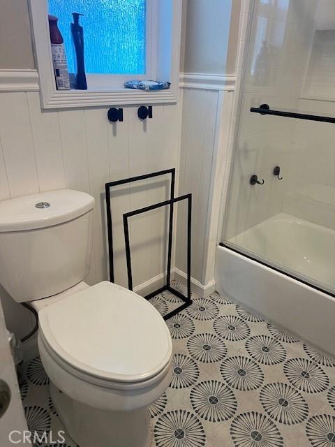 bathroom with toilet, a wainscoted wall, bath / shower combo with glass door, and tile patterned floors