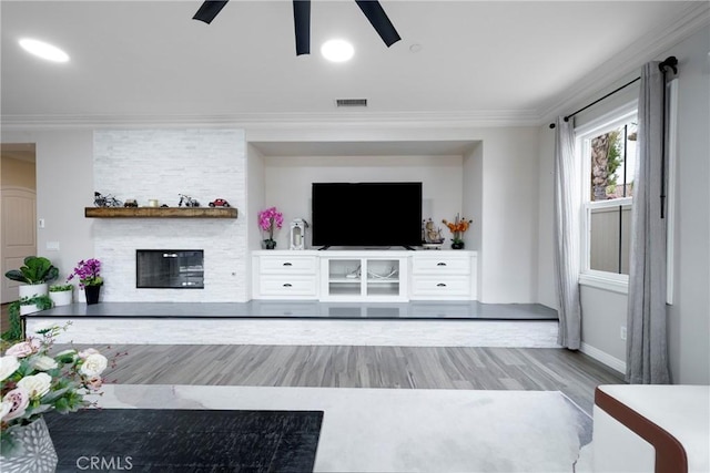 living room featuring a stone fireplace, wood finished floors, ceiling fan, ornamental molding, and visible vents