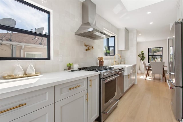 kitchen with wall chimney exhaust hood, stainless steel appliances, light wood finished floors, a sink, and tasteful backsplash