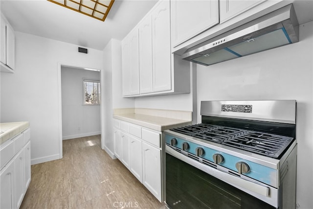 kitchen featuring tile counters, white cabinetry, under cabinet range hood, stainless steel gas range, and baseboards