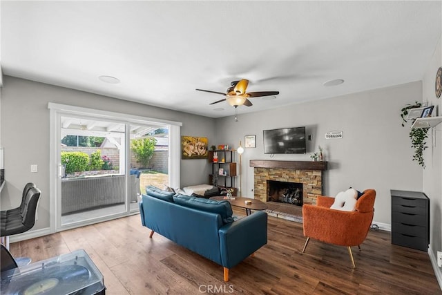 living area with baseboards, a ceiling fan, a stone fireplace, and wood finished floors