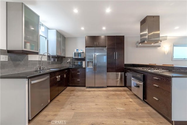 kitchen featuring light wood finished floors, stainless steel appliances, dark brown cabinets, island range hood, and a sink