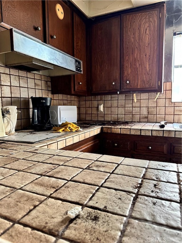 kitchen featuring under cabinet range hood, dark brown cabinetry, tile countertops, and backsplash