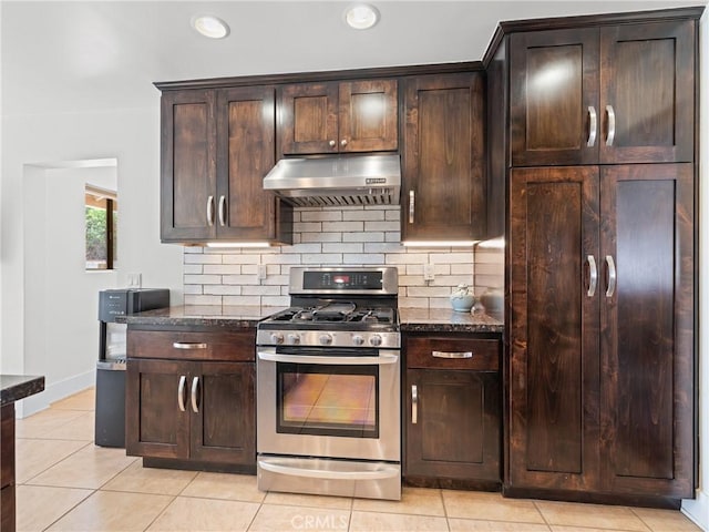 kitchen featuring tasteful backsplash, dark stone countertops, light tile patterned flooring, under cabinet range hood, and stainless steel range with gas cooktop