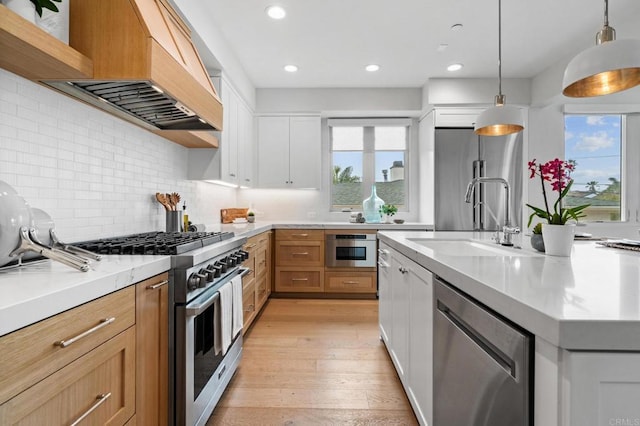 kitchen with stainless steel appliances, a sink, custom range hood, light wood-type flooring, and decorative light fixtures