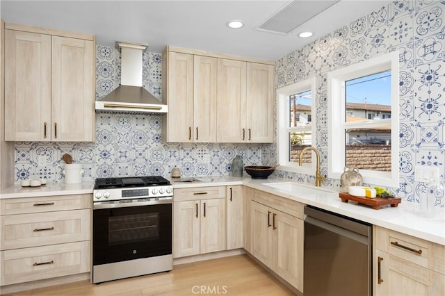 kitchen with wall chimney range hood, backsplash, appliances with stainless steel finishes, a sink, and light brown cabinetry