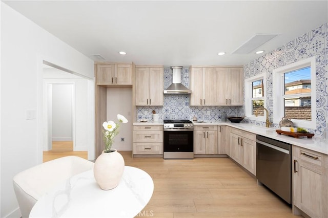 kitchen with wall chimney range hood, light countertops, stainless steel appliances, a sink, and light brown cabinetry