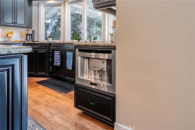 kitchen featuring light wood-type flooring, light stone counters, dishwasher, and dark cabinets