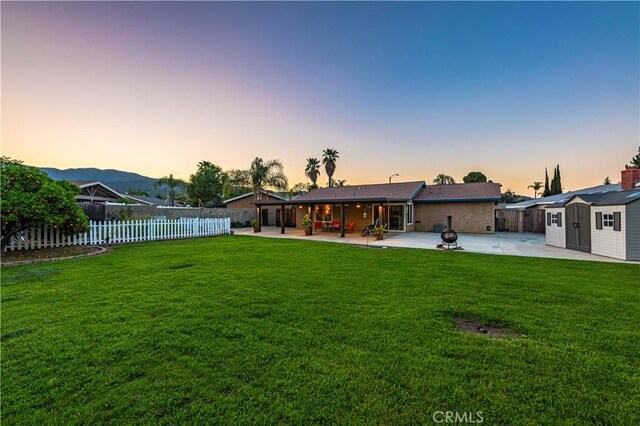 back of house at dusk featuring a storage shed, a lawn, a fenced backyard, an outdoor structure, and a patio