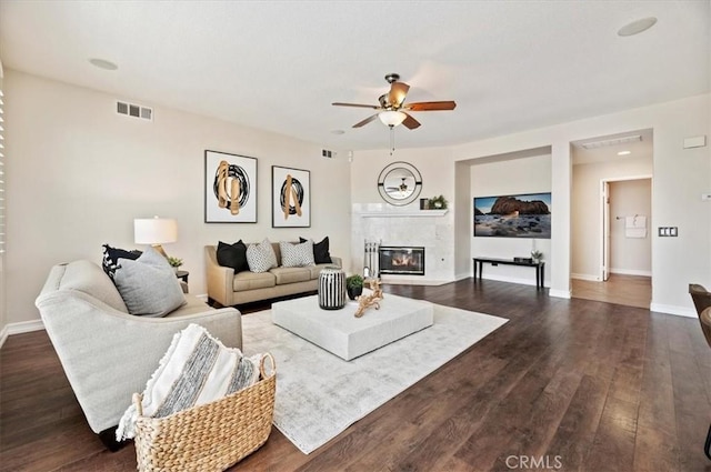 living room with dark wood finished floors, a glass covered fireplace, a ceiling fan, visible vents, and baseboards