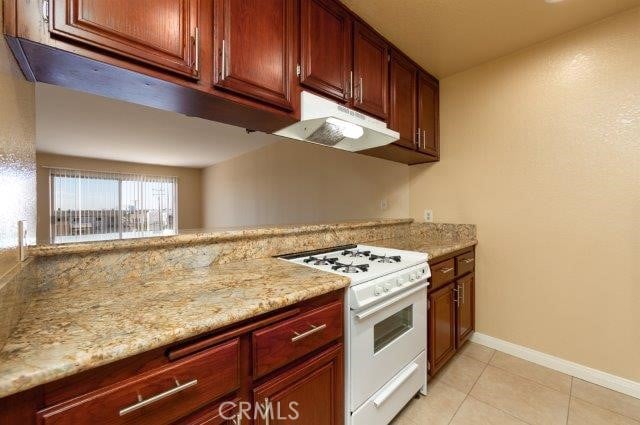 kitchen with baseboards, light tile patterned floors, white gas range, light stone countertops, and under cabinet range hood