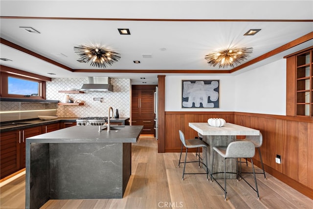 kitchen featuring dark countertops, wainscoting, a sink, wall chimney exhaust hood, and a center island with sink