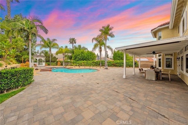 pool at dusk featuring an outdoor pool, a gazebo, a patio area, and ceiling fan