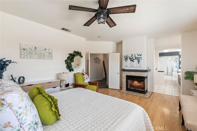 bedroom featuring visible vents, arched walkways, light wood-type flooring, a ceiling fan, and a tile fireplace