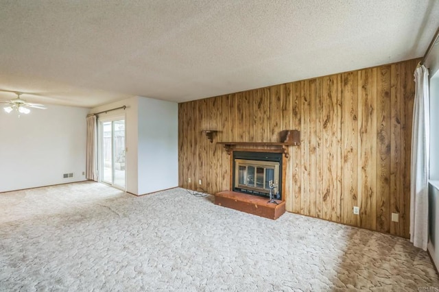 unfurnished living room with a ceiling fan, a textured ceiling, a glass covered fireplace, wood walls, and carpet flooring