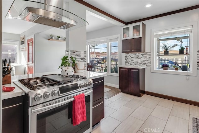 kitchen with stainless steel range with gas cooktop, glass insert cabinets, wall chimney range hood, light countertops, and decorative backsplash