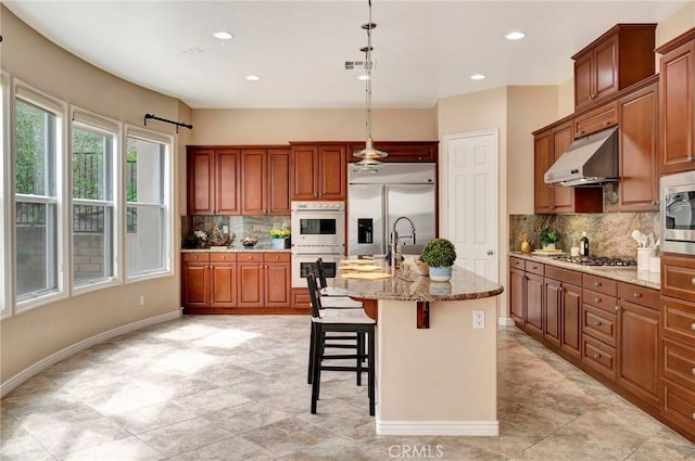 kitchen with light stone counters, under cabinet range hood, visible vents, decorative backsplash, and built in appliances