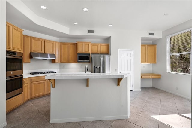 kitchen with tile counters, stainless steel appliances, under cabinet range hood, visible vents, and an island with sink