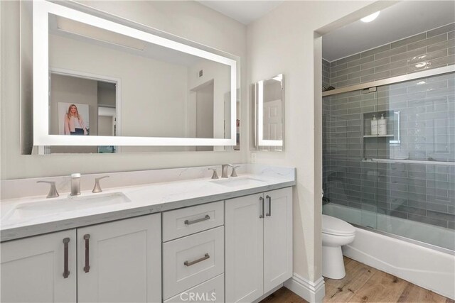 bathroom featuring a sink, shower / bath combination with glass door, double vanity, toilet, and wood finished floors