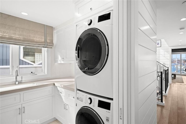 clothes washing area featuring light wood-type flooring, recessed lighting, stacked washer / dryer, cabinet space, and a sink