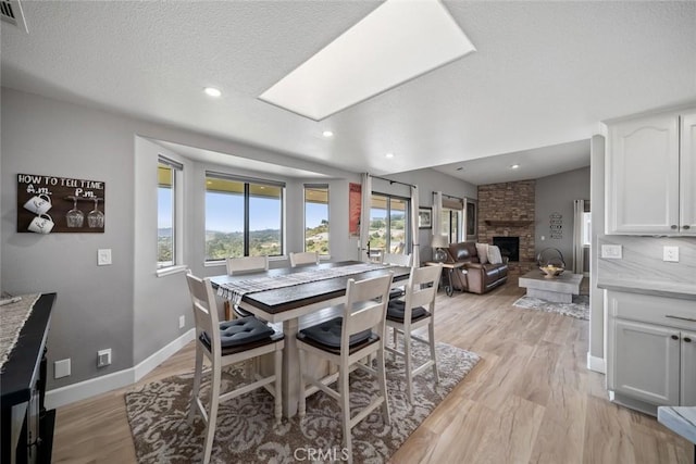dining room featuring baseboards, a stone fireplace, a skylight, a textured ceiling, and light wood-style flooring