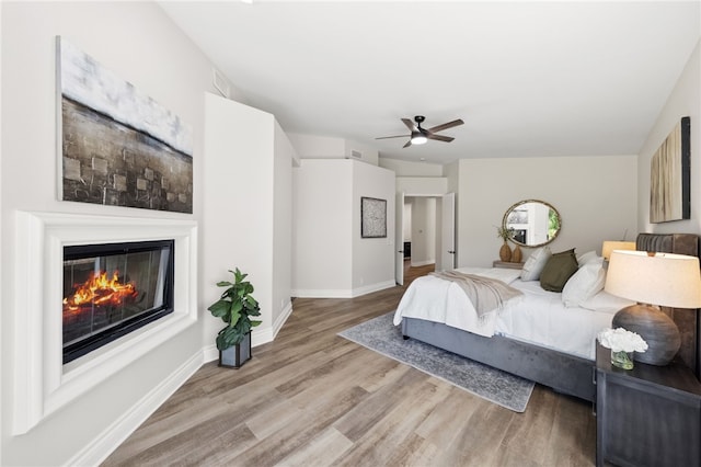 bedroom featuring wood finished floors, visible vents, baseboards, a glass covered fireplace, and ceiling fan