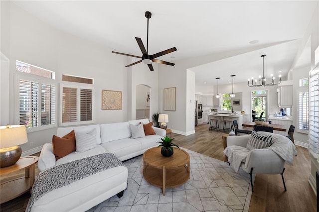 living room with a high ceiling, arched walkways, light wood-type flooring, baseboards, and ceiling fan with notable chandelier