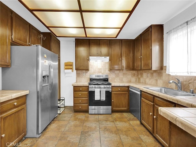 kitchen with stainless steel appliances, a sink, under cabinet range hood, backsplash, and tile counters