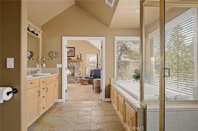 bathroom with a bath, a stone fireplace, vanity, stone tile floors, and lofted ceiling