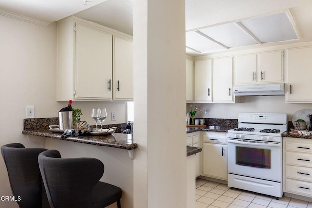 kitchen with light tile patterned floors, white gas stove, a breakfast bar, dark stone countertops, and under cabinet range hood