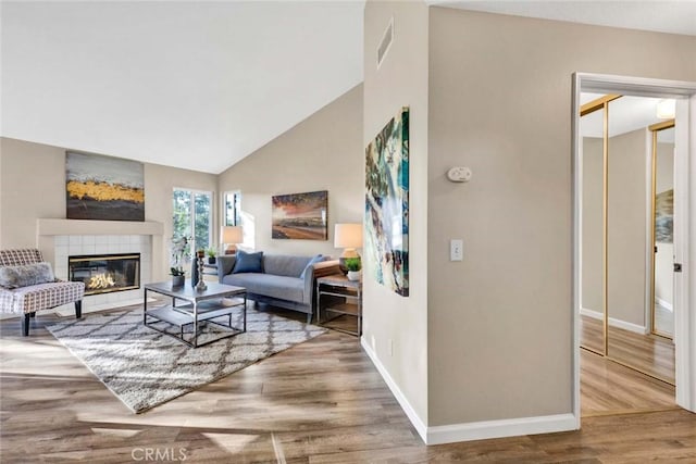 living room featuring high vaulted ceiling, wood finished floors, a tile fireplace, baseboards, and visible vents