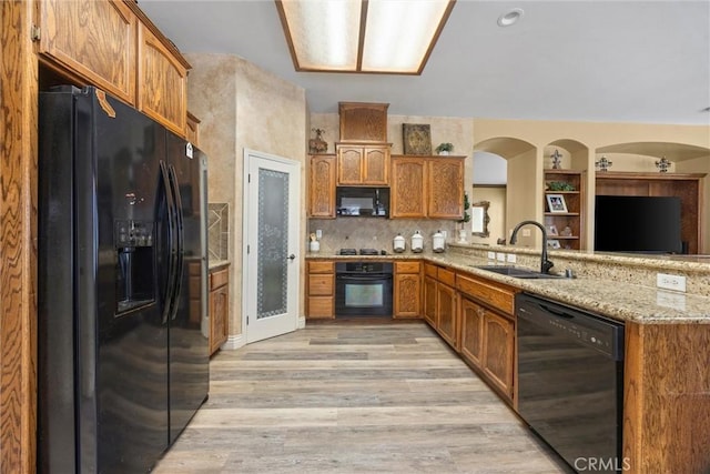 kitchen featuring light wood-type flooring, light stone counters, black appliances, brown cabinetry, and a sink