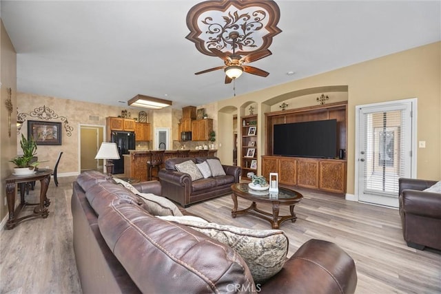 living area featuring built in features, baseboards, arched walkways, a ceiling fan, and light wood-style floors