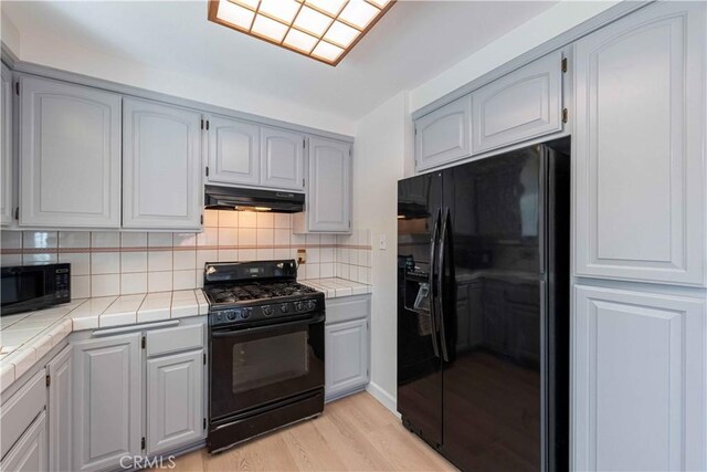kitchen featuring light wood-style flooring, under cabinet range hood, tile counters, and black appliances