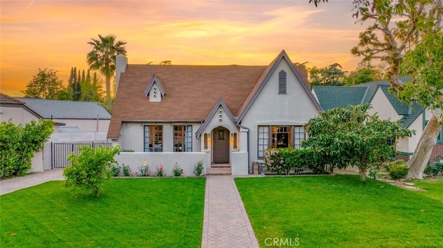 english style home with a front yard, a chimney, fence, stucco siding, and roof with shingles