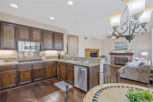 kitchen featuring appliances with stainless steel finishes, a peninsula, a brick fireplace, dark wood finished floors, and a sink