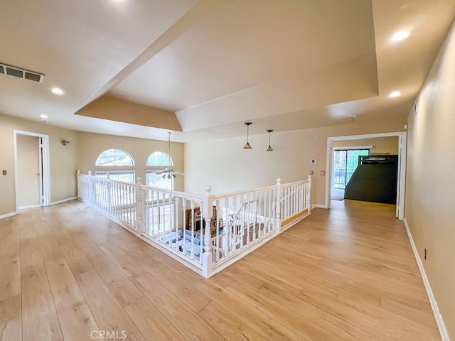 hall featuring baseboards, light wood finished floors, a tray ceiling, and an upstairs landing