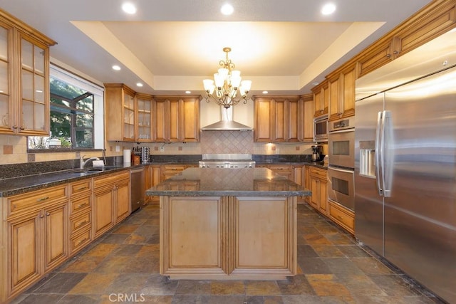 kitchen featuring wall chimney range hood, a raised ceiling, stone finish flooring, a notable chandelier, and stainless steel appliances