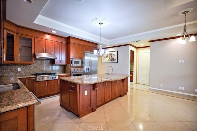kitchen with under cabinet range hood, a tray ceiling, backsplash, built in appliances, and a notable chandelier