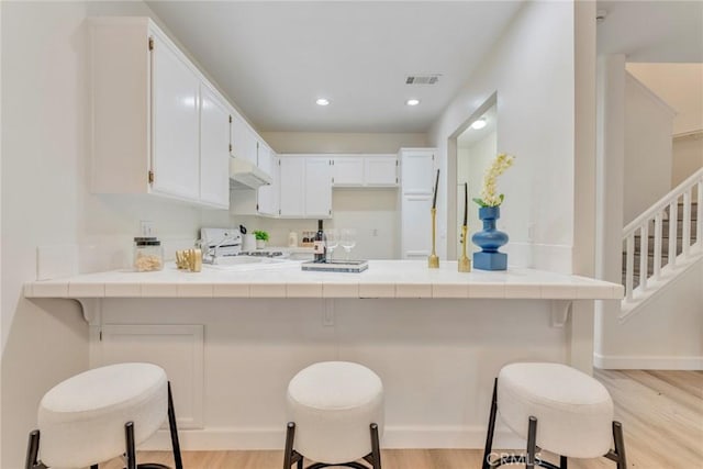 kitchen with under cabinet range hood, visible vents, tile countertops, a peninsula, and light wood-style flooring