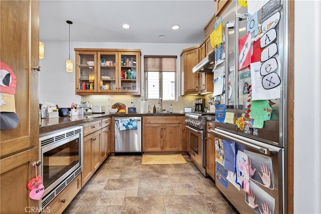 kitchen with stainless steel appliances, under cabinet range hood, brown cabinetry, stone finish floor, and dark countertops